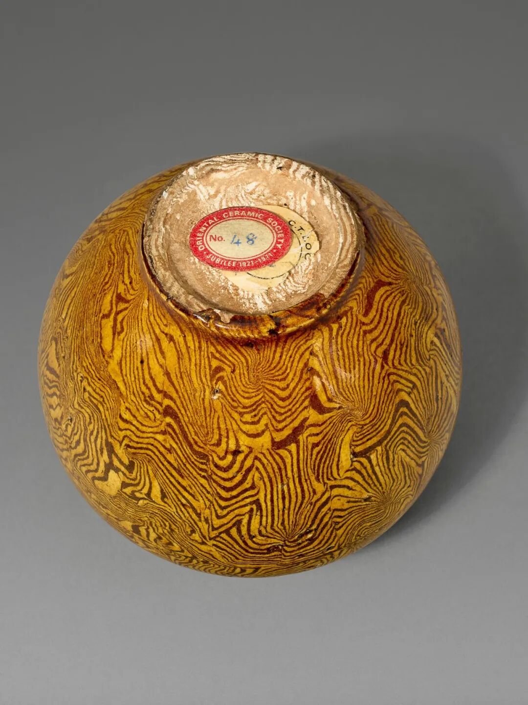 Underneath a golden-brown ceramic vessel with woodgrain pattern and red label reading Oriental Ceramic Society, No. 48 on gray background